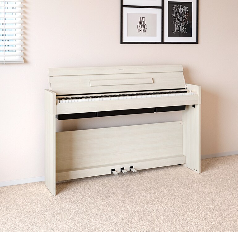 Uma sala de estar com um piano ARIUS em branco bétula (white birch), colocado junto a prateleiras de parede com candeeiros e um difusor, integrado harmoniosamente na decoração